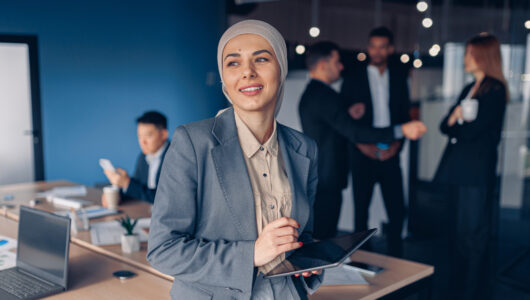Smiling muslim businesswoman in hijab working on digital tablet while standing in modern office Muslim businesswoman in hijab looking at side working on digital tablet on colleagues background